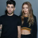 Zayn Malik i Gigi Hadid pozują razem, oboje ubrani na czarno. Zayn ma tatuaże na ramionach, a Gigi odsłania brzuch w krótkim topie.