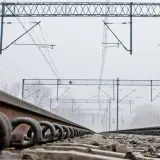 Torowisko kolejowe z bliska, z widocznymi śrubami mocującymi szyny. W tle sieć trakcyjna i mglisty krajobraz.