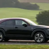 Czarna Mazda MX-30 z bordowym dachem sunie po drodze wśród zielonych wzgórz.