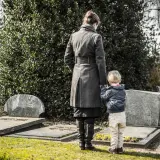 Kobieta z dzieckiem na cmentarzu. Dziecko trzyma matkę za rękę, wspólnie przeżywają żałobę po babci.