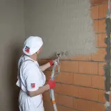 Pracownik w białym stroju nakłada tynki cementowo wapienne na ścianę z pustaków ceramicznych za pomocą agregatu.