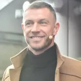 Uśmiechnięty Dariusz Pachut w beżowym płaszczu i czarnym golfie, z mikrofonem przy uchu. Może opowiada o swojej żonie.