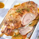 Gigot d'agneau désossé au four, doré et juteux, garni de romarin et d'ail. Servi avec sa sauce dans un plat blanc.