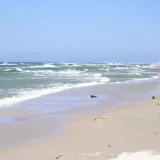 Plaża w Rowach, z falującym morzem i piaszczystym brzegiem. W oddali widać wydmy i nielicznych spacerowiczów.