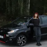 En kvinna med rött hår lutar sig mot en svart Hyundai i10. Bilen är redo för ett test.