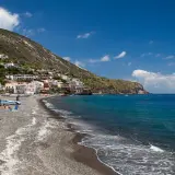 Vue d'une plage de galets sur l'île de Lipari, avec des maisons colorées accrochées à la colline et des bateaux dans la mer d'un bleu profond.