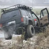 Czarny Jeep z bagażnikiem dachowym pokonuje trudny teren, to prawdziwe off road warszawa! Dym z opon i otwarta drzwi dodają dynamiki.