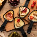 Raclette party with melted cheese, bacon, mushrooms, and tomatoes. Discover par quoi remplacer le fromage à raclette pour varier les plaisirs !