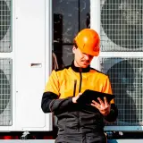 Technik w pomarańczowym kasku sprawdza dane na tablecie przy jednostkach zewnętrznych pompy ciepła, planując dofinansowanie.