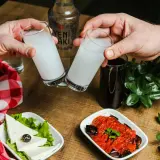 Dwie szklanki z mleczną wódką turecką (rakı) unoszą się do toastu. Na stole ser biały, oliwka i sos pomidorowy z bakłażanem.