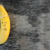 Banan z namalowaną buźką i kciukiem w górę, ubrany w **kondom**. To zabawne przypomnienie o bezpieczeństwie.