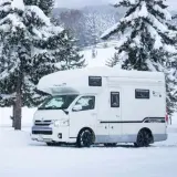 Camping car japonais blanc garé dans la neige, entouré de sapins enneigés. Des remontées mécaniques sont visibles en arrière-plan.