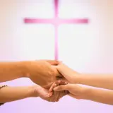 Para młoda trzyma się za ręce, przygotowując się do nauk przedmałżeńskich. W tle różowy krzyż.
