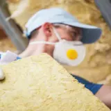 Operaio con mascherina e guanti maneggia pannelli isolanti, potenzialmente lana di roccia cancerogena, in un sottotetto.