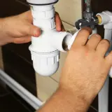 Ręce majsterkowicza składają biały syfon pod zlewem, przygotowując naprawę rury pcv.