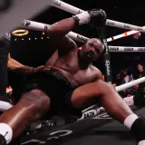 Derrick Chisora klęczy na macie, wyczerpany po walce. W tle widać publiczność i ring bokserski.
