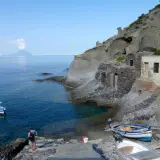 Sur l'île Salina, un homme amarre un bateau près de vieilles maisons de pêcheurs en pierre, avec une île volcanique au loin.