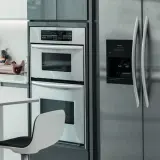 Nowoczesna kuchnia z lodówką obok piekarnika w zabudowie. Dwa stylowe krzesła barowe stoją przy blacie.