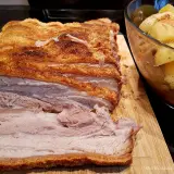 Ein butterzarter Schweinebauch mit knuspriger Kruste liegt auf einem Holzbrett, daneben eine Schüssel mit Bratkartoffeln und Gemüse.