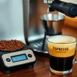 Waga pokazuje 18g ziaren kawy, idealna ilość na filiżankę espresso. Obok stoi szklanka z aromatycznym napojem.