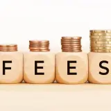 Cztery drewniane kostki z literami F, E, E, S tworzą napis FEES. Na każdej kostce stos monet, rosnący od lewej do prawej.