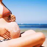 Brzuszek w ciąży na plaży, posmarowany kremem z filtrem w kształcie słońca. Bezpieczne opalanie w ciąży to podstawa.