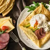 Złożone crepes z jajkiem w koszulce, awokado, szynką i serem. Obok stos naleśników, pomidorki i rukola.