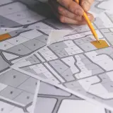 Rysunek geodezyjny z zaznaczonymi działkami. Dłoń z ołówkiem planuje połączenie a scalenie działek.