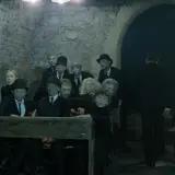 Postacie z "Umarłej klasy" Tadeusza Kantora, w tym uczniowie i nauczyciele, siedzą w ponurej scenerii.
