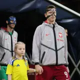 Polscy piłkarze, w tym Robert Lewandowski, wychodzą na murawę z dziećmi. Jeden z zawodników ma niebieską maskę.