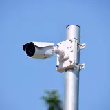 Biała kamera zewnętrzna zamontowana na metalowym słupku, idealna do monitorowania otoczenia. Dowiedz się, jak zamontować kamerę zewnętrzną.