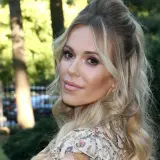 Doda IQ, blondynka w kwiecistej sukience, z burzą loków, uśmiecha się do obiektywu na tle zieleni.