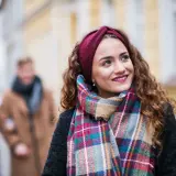 Uśmiechnięta dziewczyna w bordowej opasce i kolorowym szalu, idealnym do płaszcza w kratę. W tle para spacerująca ulicą.
