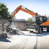 Pomarańczowa koparka Doosan DX150W pracuje na placu budowy w Poznaniu, rozbierając betonowe ściany.