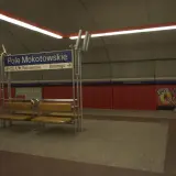 Na stacji metra Pole Mokotowskie stoi mężczyzna obok ławki. W tle niebieska tablica z nazwami ulic i czerwone panele.
