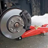 Hamulec samochodowy z tarczą i zaciskiem, gotowy do wymiany. Czerwony podnośnik hydrauliczny, jak jarzmo, czeka na swoją kolej.