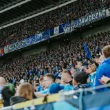 Tłum kibiców na stadionie, w niebieskich barwach, z transparentami "Jaworzno", "Mikołów", "Chorzów". Wszyscy w zgodzie dopingują swoją drużynę.