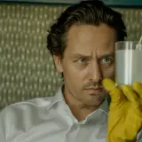 Ein Mann mit blauen Augen und gelben Handschuhen hält ein Glas mit weißer Flüssigkeit. Er ist Teil der "Achtsam Morden Reihe".