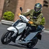 Hombre en scooter Peugeot Tweet 125, con casco y chaqueta verde, circulando por una calle.