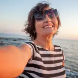 Uśmiechnięta kobieta w okularach przeciwsłonecznych robi selfie na plaży o zachodzie słońca.