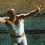 Aria dla atlety obsada: cyrkowy artysta w błyszczącym stroju z wąsami, z rękami uniesionymi w geście powitania.
