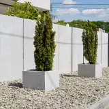 Nowoczesne ogrodzenie od drogi powiatowej z betonu, z dwoma stożkowymi drzewkami w kwadratowych donicach na tle żwirowego podjazdu.