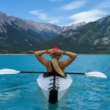 Frau im Kajak auf türkisfarbenem See, Berge im Hintergrund. Ein Symbol für das Erreichen von finanzieller Freiheit und ein Leben voller Abenteuer.
