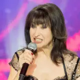 Krystyna Giżowska śpiewa do mikrofonu, w czarnej, błyszczącej sukience. Wiele osób zastanawia się, ile lat ma Krystyna Giżowska.