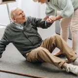Älterer Mann sitzt auf dem Boden, wird von einer Frau gestützt. Ein Stock liegt daneben. Dies illustriert die Notwendigkeit von Sturzprophylaxe.