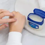 Dłoń rozsmarowuje na skórze nawilżający krem z wazeliną. Obok stoi otwarty słoiczek z produktem.