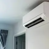 Biała jednostka klimatyzacyjna zamontowana na jasnej ścianie, obok drzwi i okna z zasłonami.