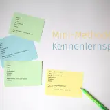 Bunte Zettel mit Fragen für ein Kennenlernspiel liegen auf weißem Grund. Ein grüner Stift liegt daneben.