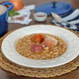 Plato de judías blancas con chorizo y tocino, servido en una vajilla blanca sobre una esterilla de mimbre.
