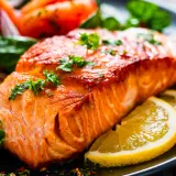 Un succulento filetto di salmone cotto alla perfezione, guarnito con prezzemolo fresco e accompagnato da spicchi di limone e pomodorini.